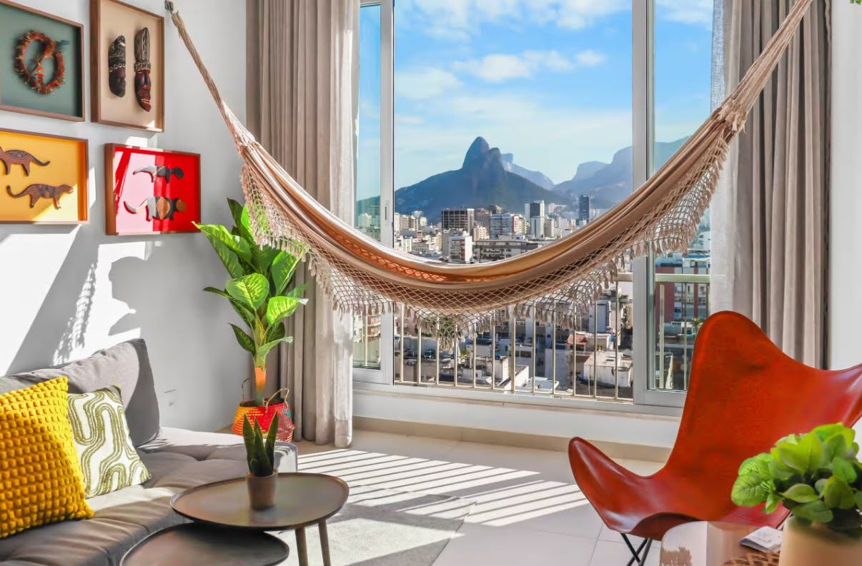 A living room with a fringed hammock hung across a large window, a grey sofa with yellow cushions, an orange leather butterfly chair and framed wall pieces, with city rooftops and a twin-peaked mountain outside.