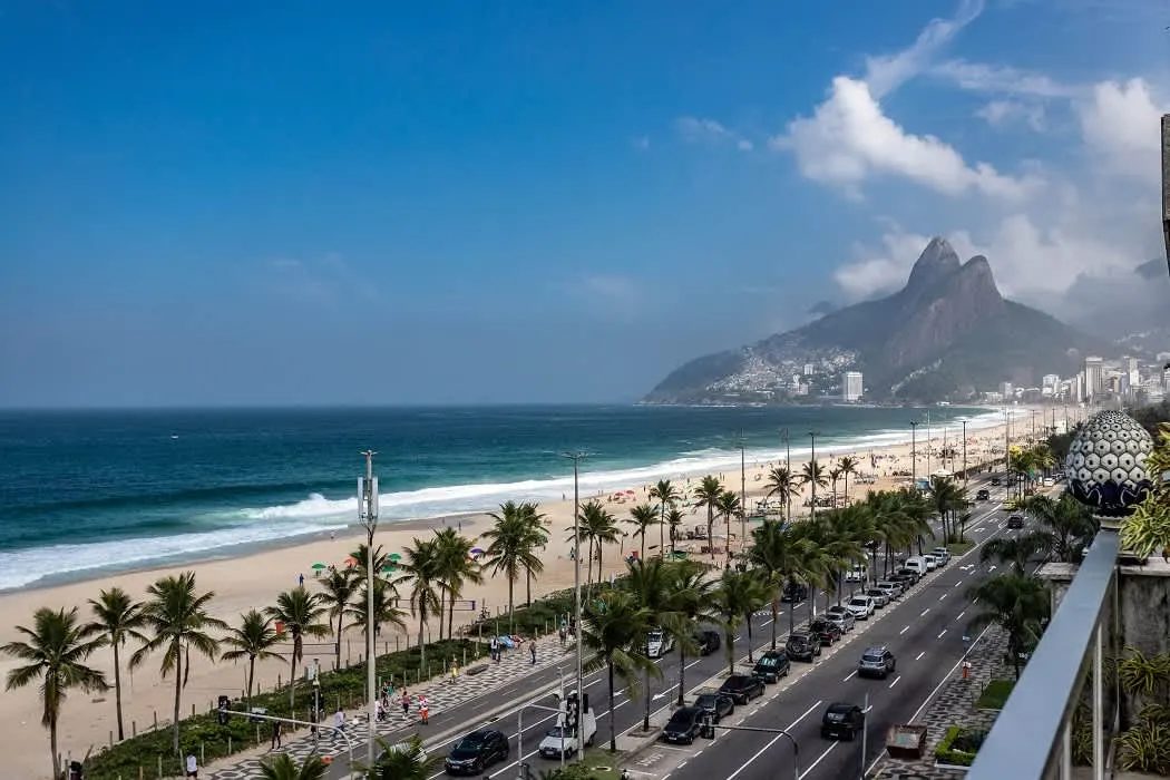 A long sandy beach with breaking waves beside a palm-lined avenue with cars, seen from a high terrace, with a twin-peaked mountain and a hillside of small houses at the far end.
