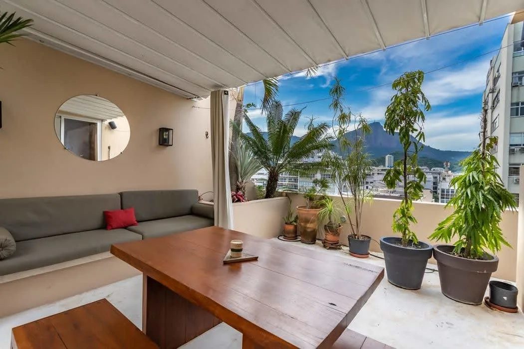 A covered roof terrace with a built-in grey sofa, a wooden table and benches, a round mirror and potted plants, looking over rooftops to a mountain under a blue sky with clouds.