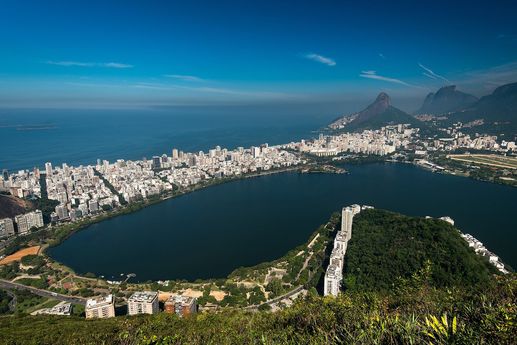 A high view over a dark blue lagoon ringed by parkland and apartment buildings, with a strip of dense city between the lagoon and the ocean and steep rocky peaks to the right.