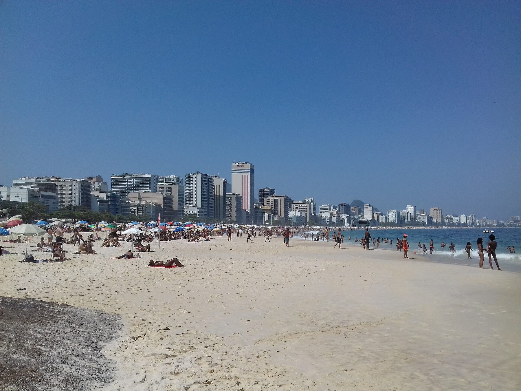A wide sandy beach with sunbathers, parasols and people at the water's edge, and a long line of apartment buildings and a striped tower marked Marina along the shore under a clear sky.