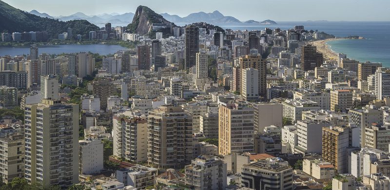A dense spread of apartment towers seen from above, with a lagoon on the left, a steep rock hill in the middle distance and a curving sandy beach and the ocean on the right.