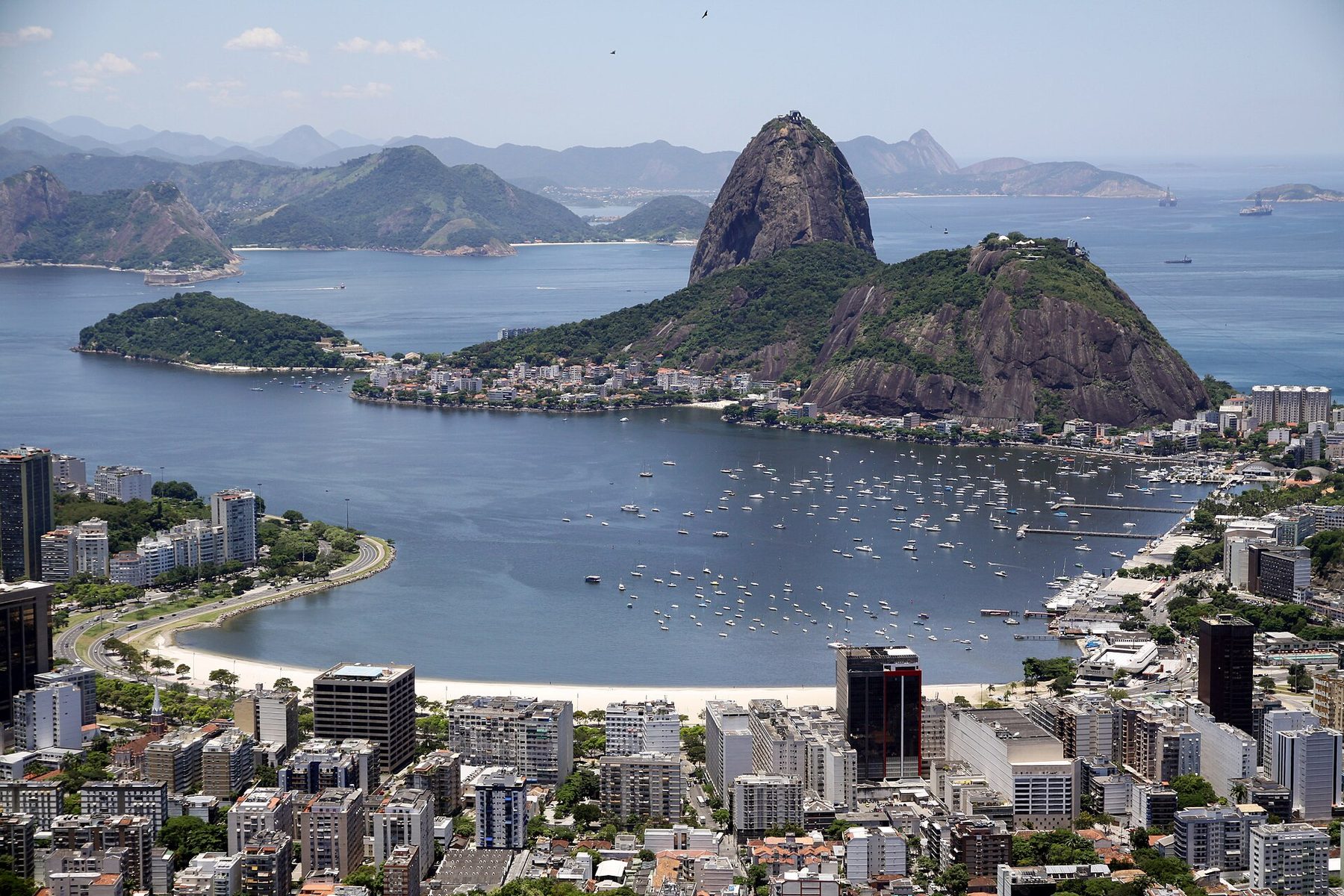 A round bay dotted with moored sailing boats, with a tall bare rock peak and a lower green hill on a peninsula behind it, city buildings in the foreground and hazy hills across the water.