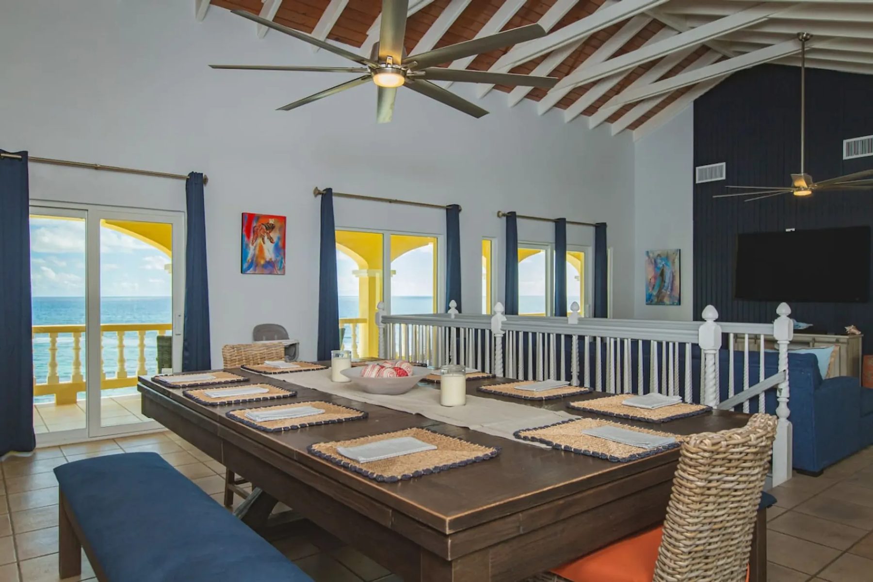 A long dark wooden dining table laid with ten woven place mats, a bench and wicker chairs, under a vaulted ceiling with white beams and a large fan; glass doors open to a yellow veranda and the sea.