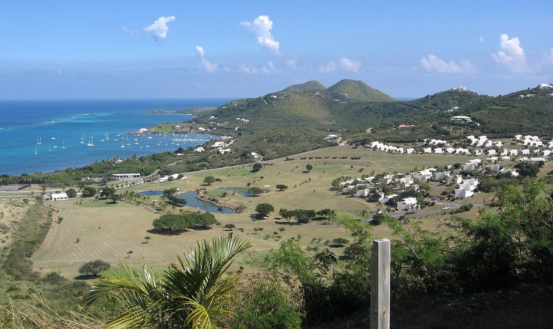 View from a hill over a golf course with ponds and white low-rise buildings to a bay of turquoise water with many moored sailboats, and rounded green hills along the coast beyond.