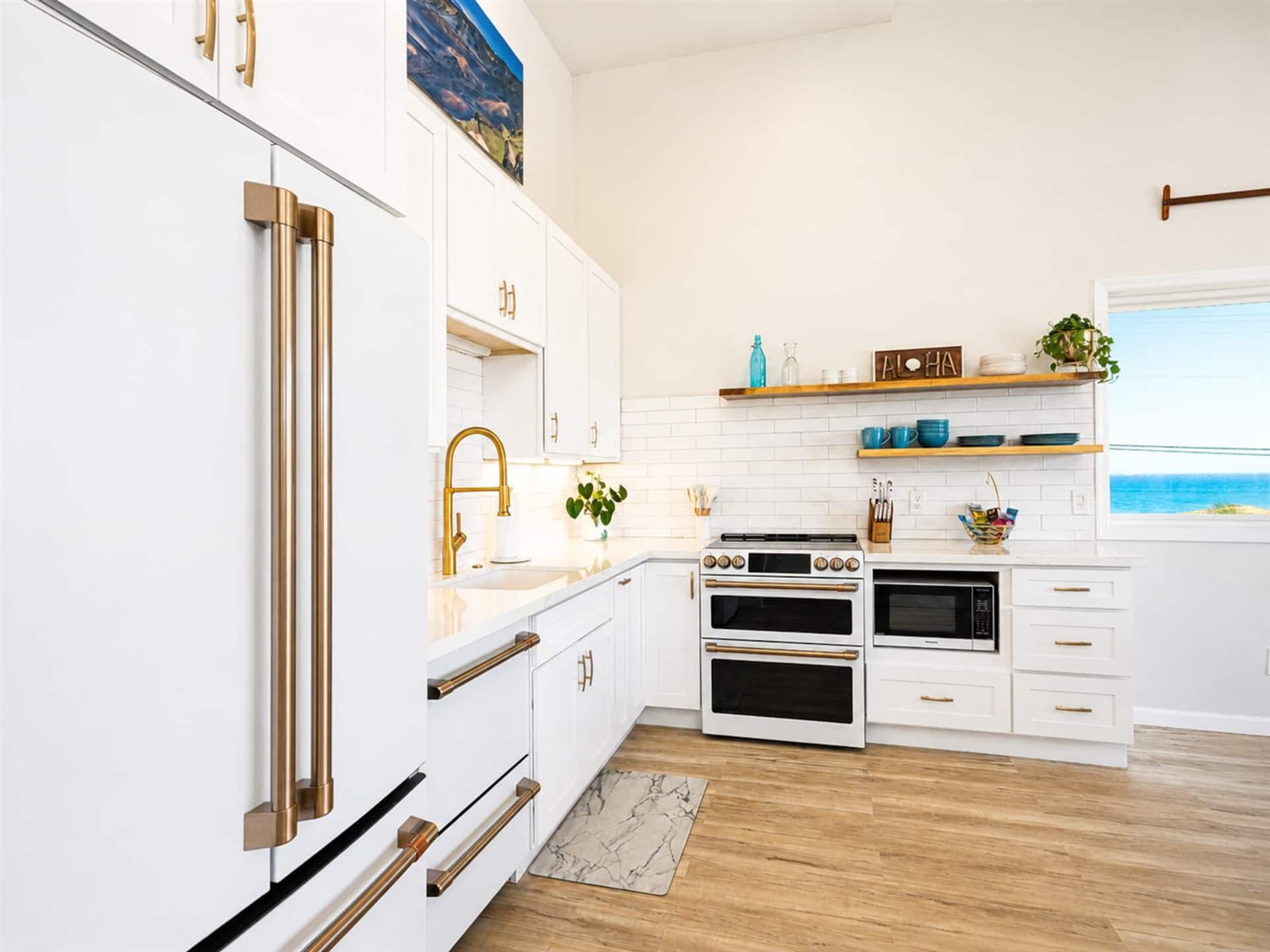 White kitchen with a French-door refrigerator with brass handles, a double-oven range, white subway tile, open wooden shelves holding blue dishes and an Aloha sign, and a window showing the sea.