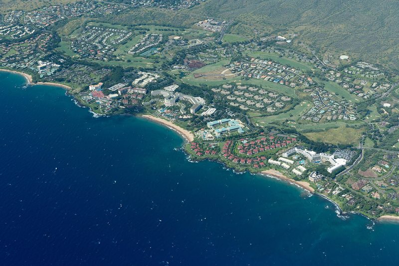 Aerial view of a coastline with several large resort hotels, pools, red-roofed condominiums, golf fairways and housing behind crescent sand beaches and dark blue sea.