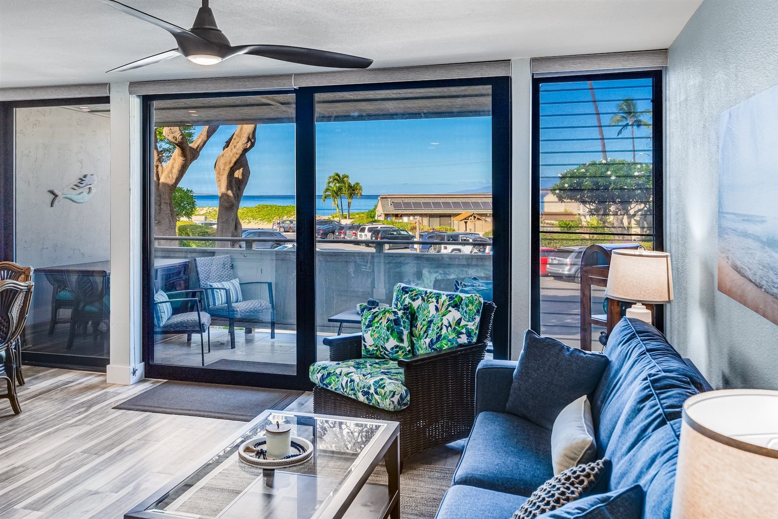 Living room with a blue sofa, a leaf-print wicker armchair and a glass coffee table, facing sliding glass doors to a balcony; beyond are parked cars, a low building with solar panels, dunes and the sea.