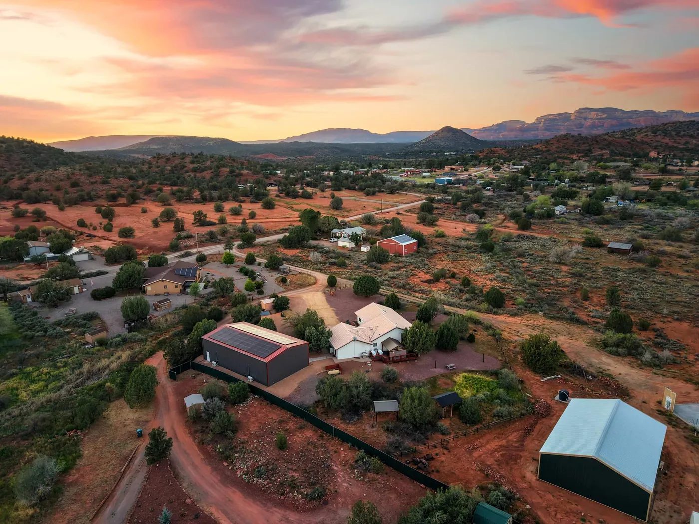 Drone view at sunset over a scattered rural neighbourhood on red soil: in the foreground a dark metal building with solar panels on its roof stands beside a white house; hills and distant cliffs line the horizon.