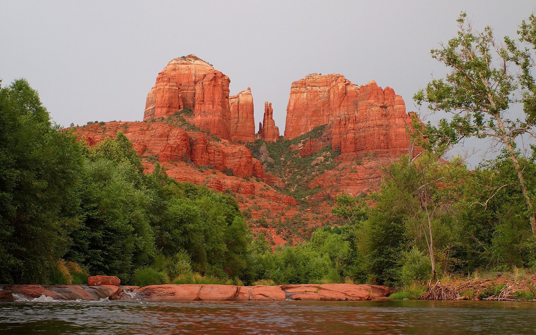 Two broad red sandstone buttes with thin spires between them rise above green trees; a creek flows over flat red rock ledges in the foreground under a grey sky.