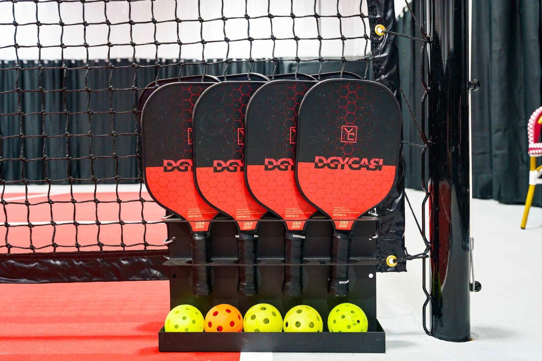 Four red-and-black pickleball paddles stand in a black rack beside the net post, with four yellow balls and one orange ball in a tray beneath them, on the orange court.