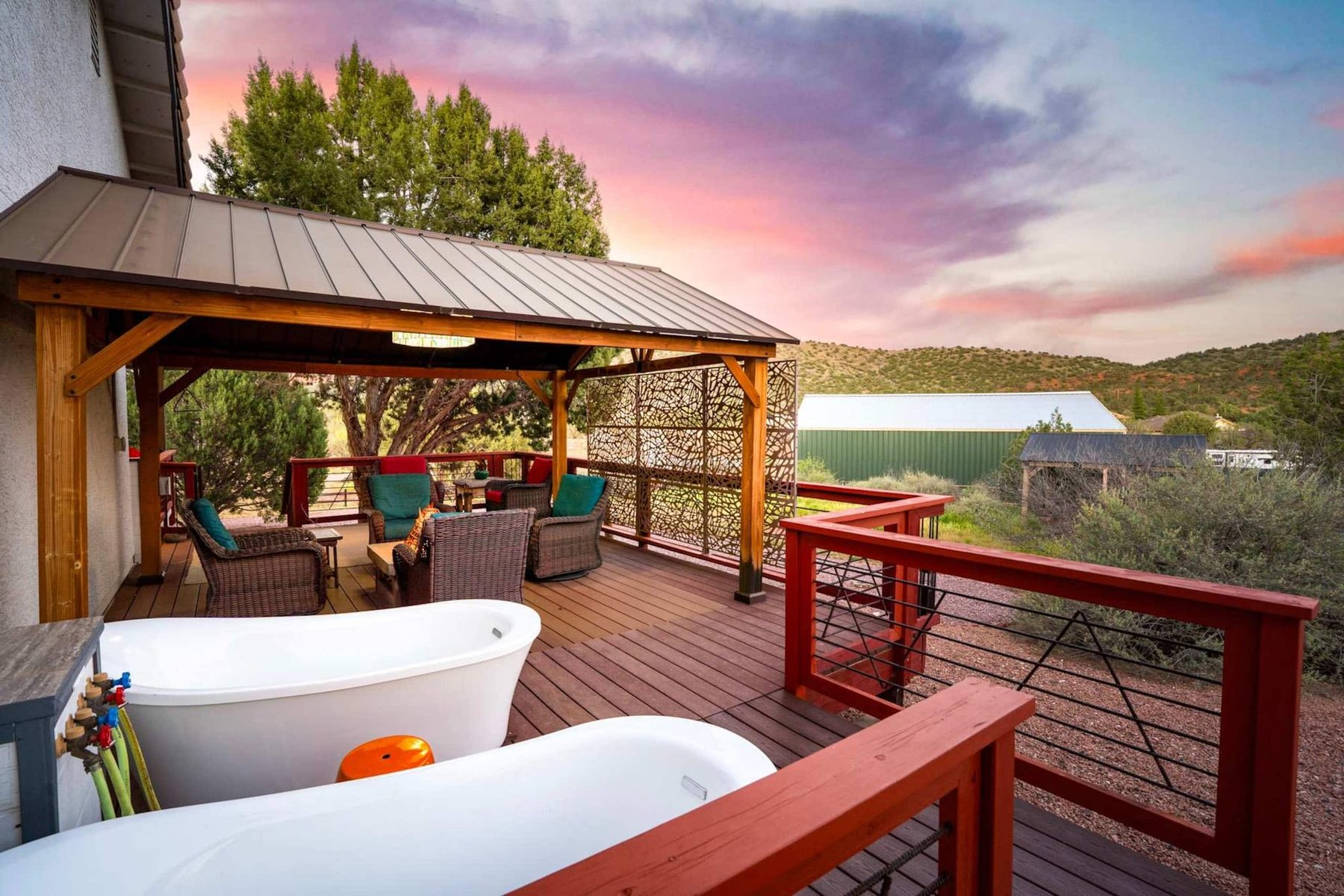 Two white freestanding bathtubs on a wooden deck with red railings, beside a timber pergola with wicker chairs and a patterned metal screen; juniper trees, a green metal building and low hills lie beyond under a pink dusk sky.