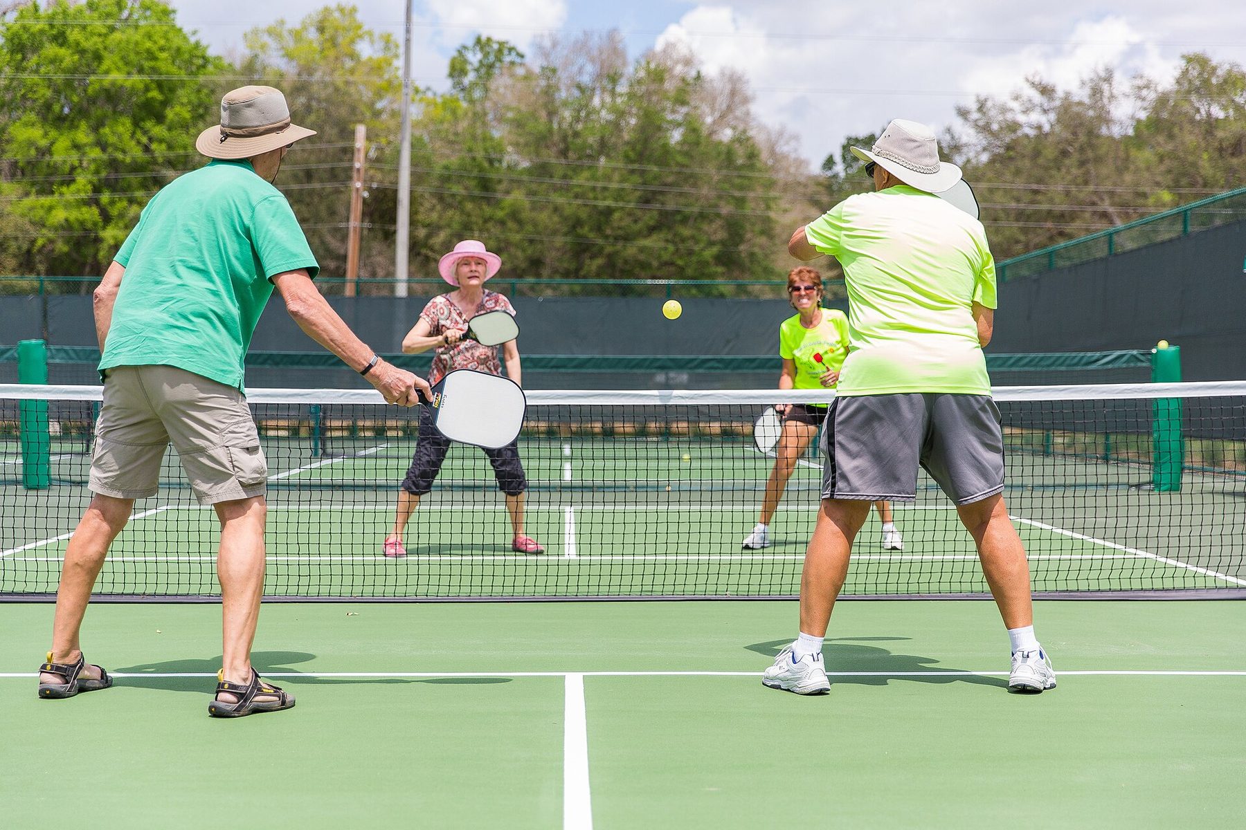 Four older players in sun hats and bright shirts play doubles on a green outdoor pickleball court, two on each side of the net, with a yellow ball in the air.