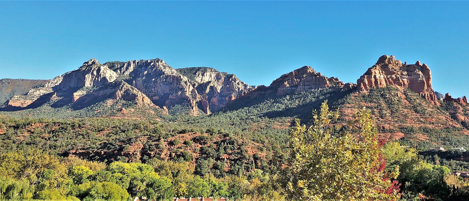 A long ridge of red and cream sandstone buttes rising above green desert scrub and trees at Sedona, Arizona, under a blue sky.