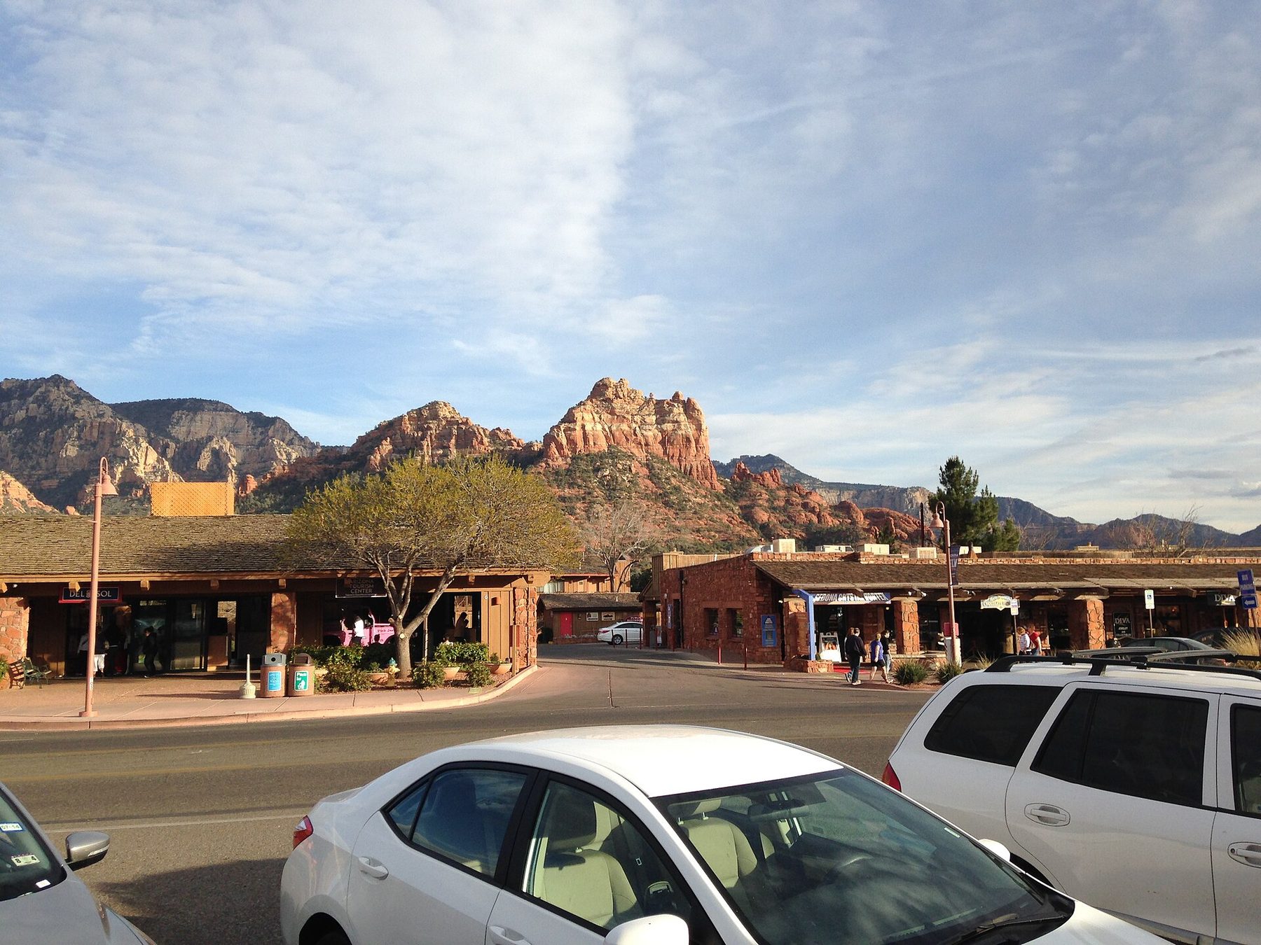 A street of low wood-and-stone shop buildings with covered walkways, a few pedestrians and parked white cars in the foreground; red rock buttes and cliffs rise behind under a streaky sky.