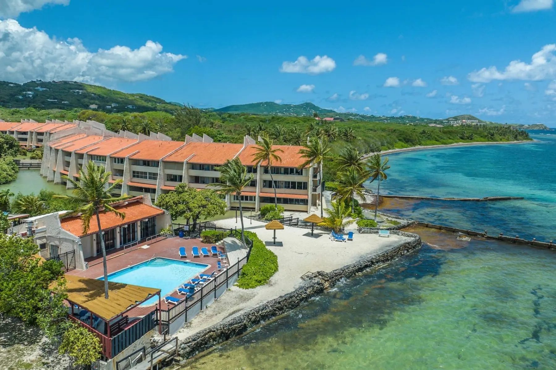 An aerial view of a three-storey condominium building with orange tile roofs beside a shared pool, a small walled beach with thatched umbrellas and shallow green water along a wooded shore.