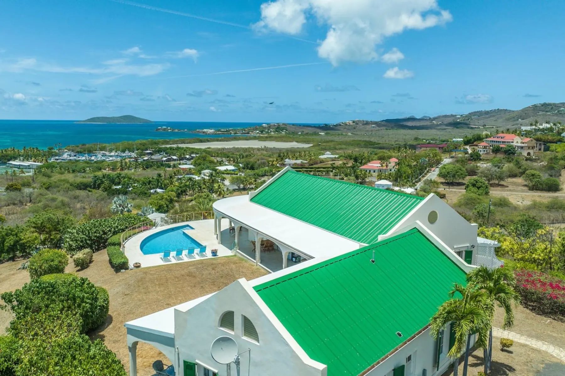 An aerial view of a white house with bright green metal roofs, an arched veranda and a curved pool on a dry hillside, with a marina, a bay and a small island on the horizon.
