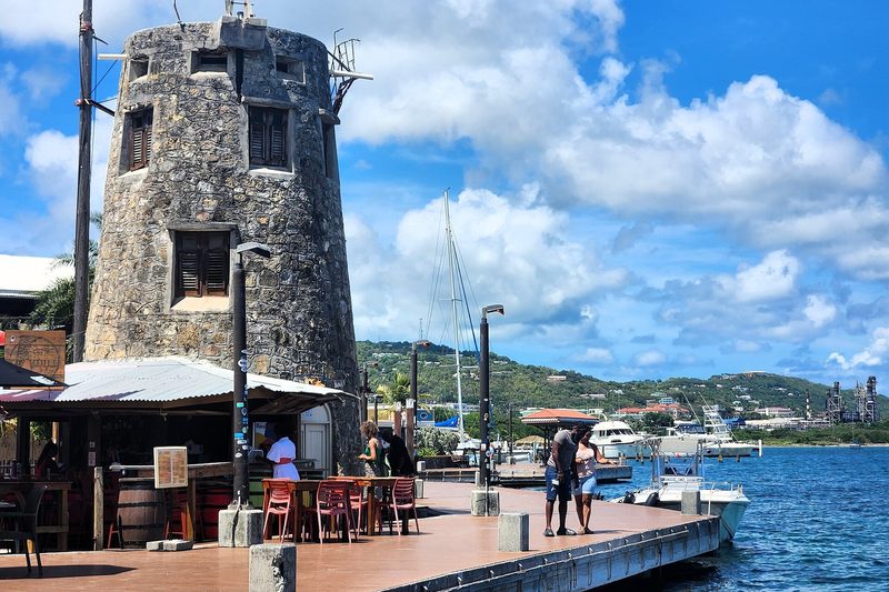 A round stone windmill tower beside a wooden boardwalk with a covered bar and tables, boats tied alongside and yachts in the harbor behind, under a blue sky with clouds.