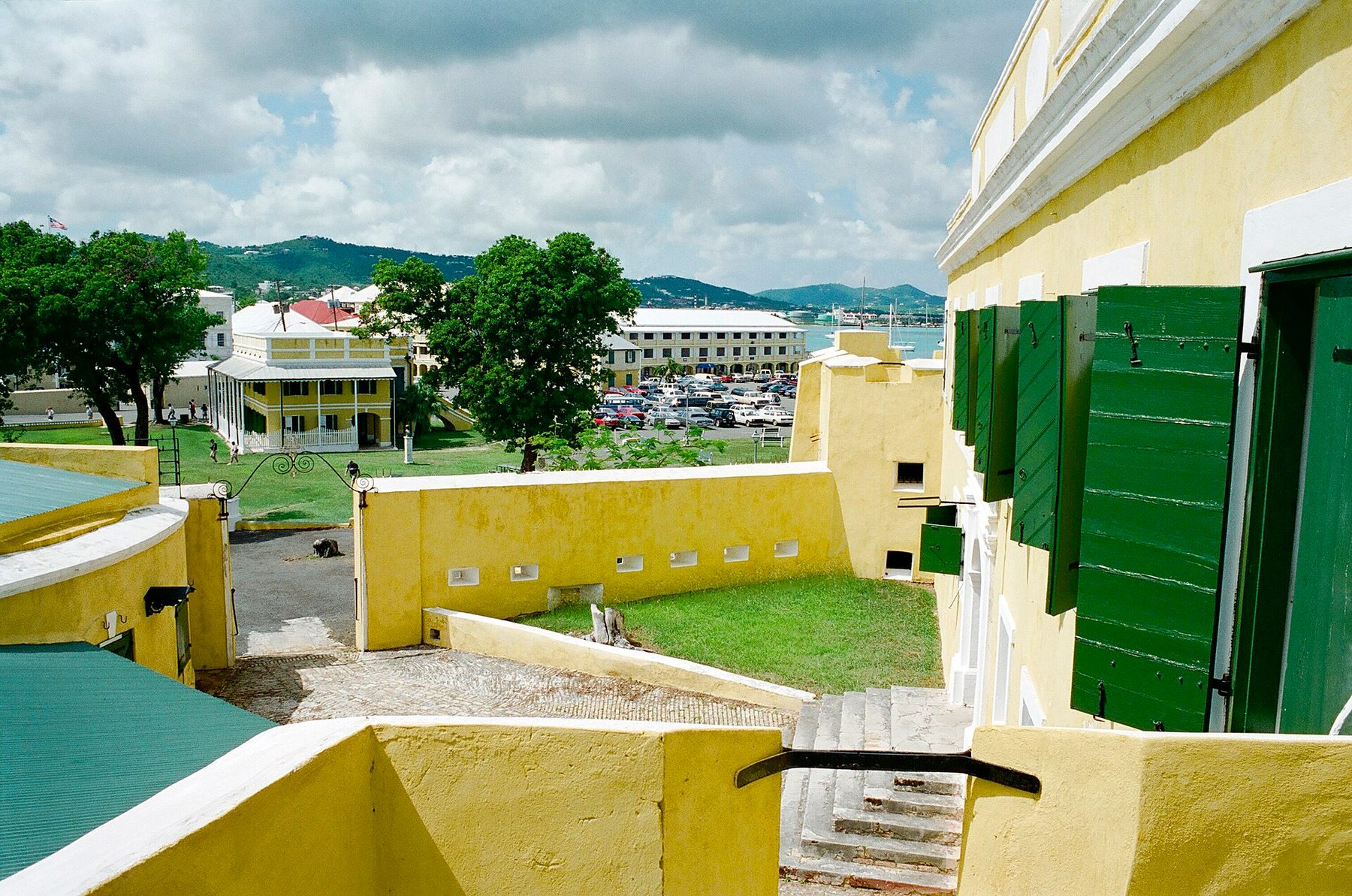 The yellow walls, green shutters and cobbled ramp of an old fort, looking out over a lawn to yellow colonial buildings, a car park and a harbour backed by green hills.