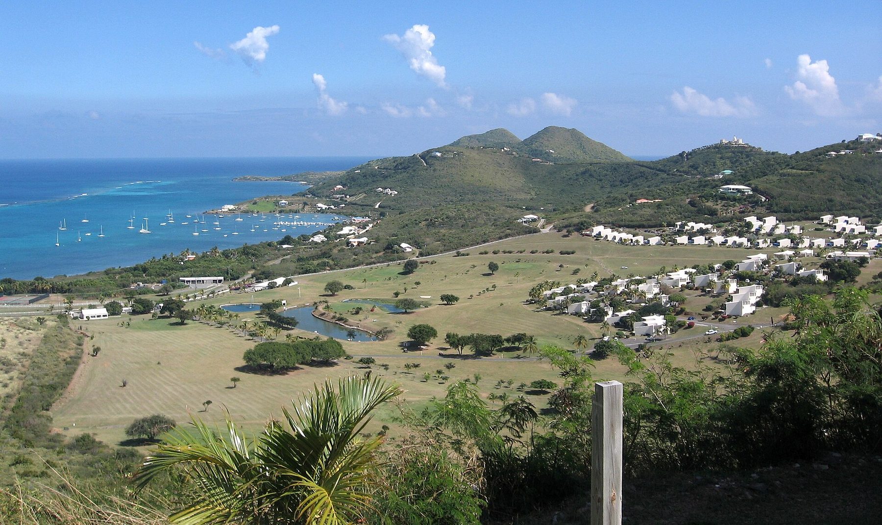 A view from a hillside over a golf course with ponds and clusters of white buildings to a bay full of anchored sailing boats, a reef line and two rounded green hills.