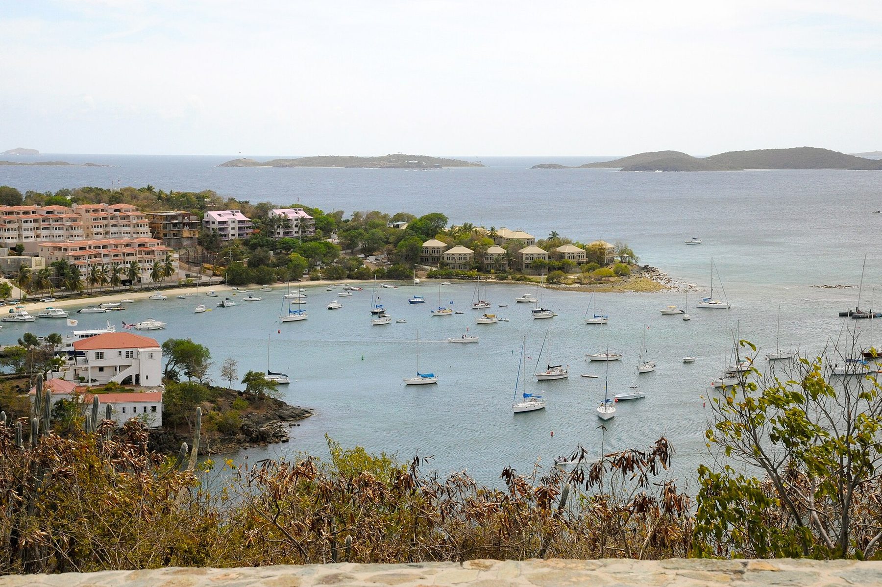 A sheltered bay crowded with moored sailing boats, with terracotta-roofed buildings along the shore and low islands on the horizon, seen from a hillside of dry scrub and cactus.