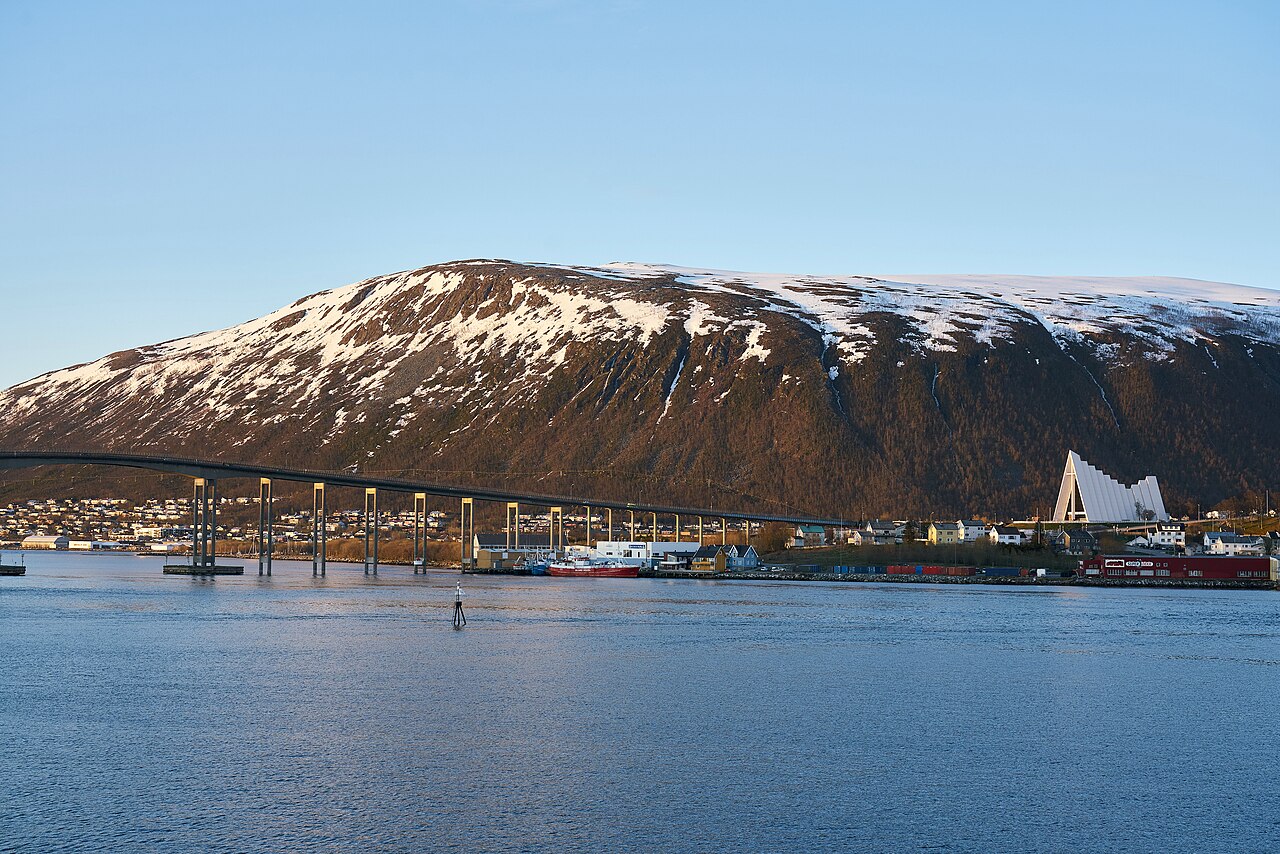 A long road bridge crosses calm blue water toward a white triangular church at the foot of a snow-streaked mountain.