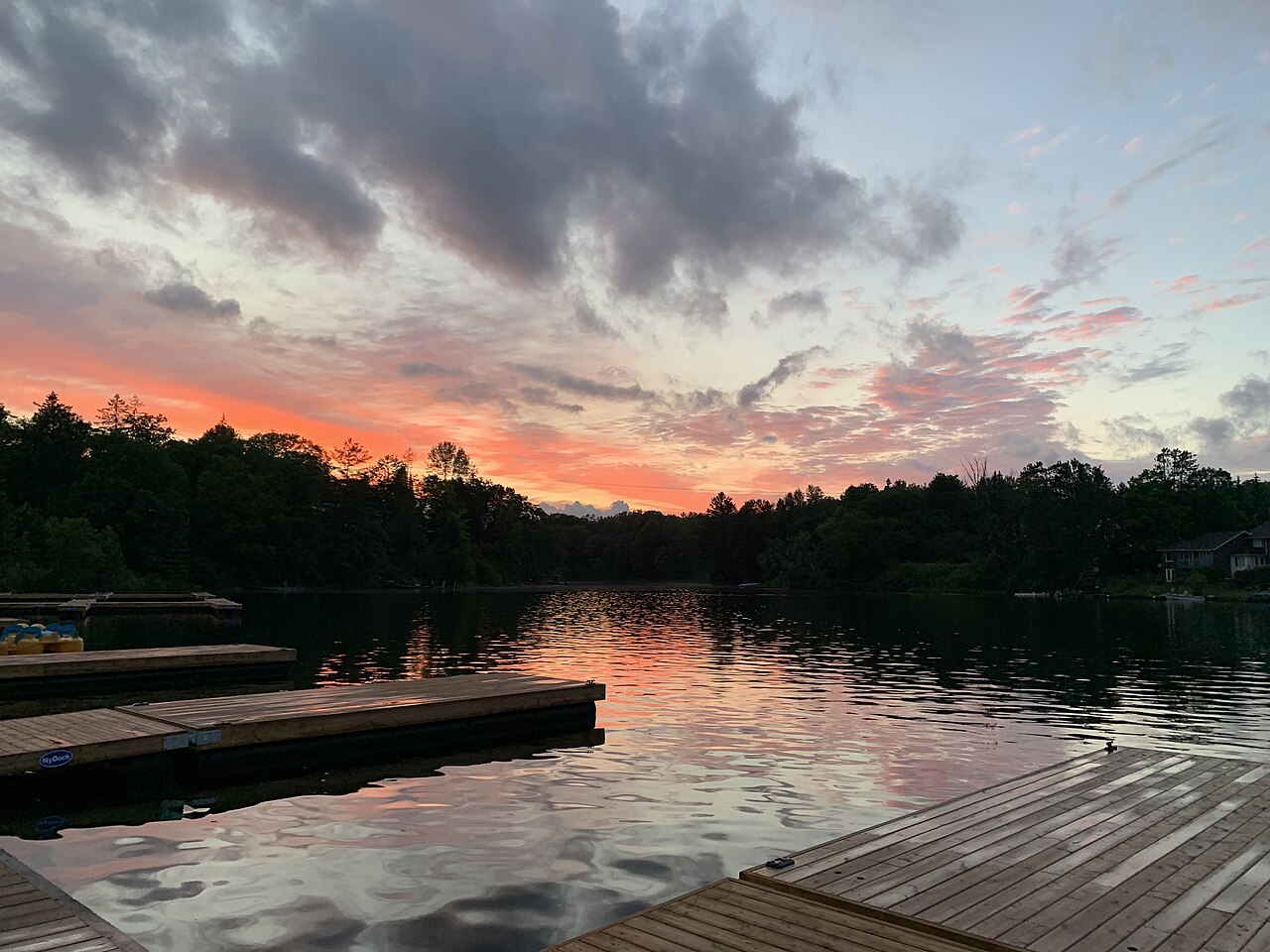 Pink and orange clouds reflected in a still lake edged by dark forest, with wooden docks in the foreground.