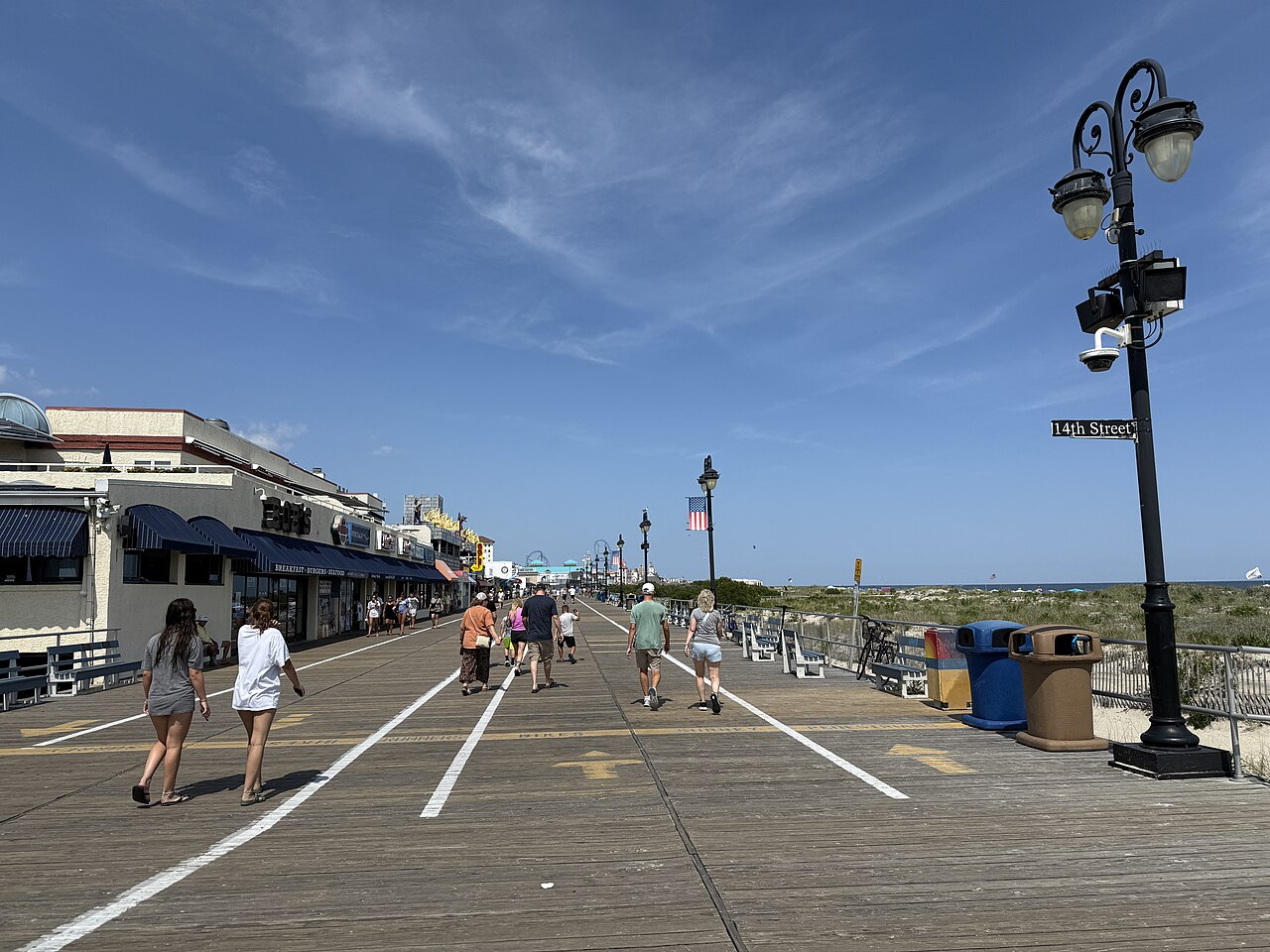 A wide wooden boardwalk with painted lanes runs past low shops toward the horizon, with dunes on the right.