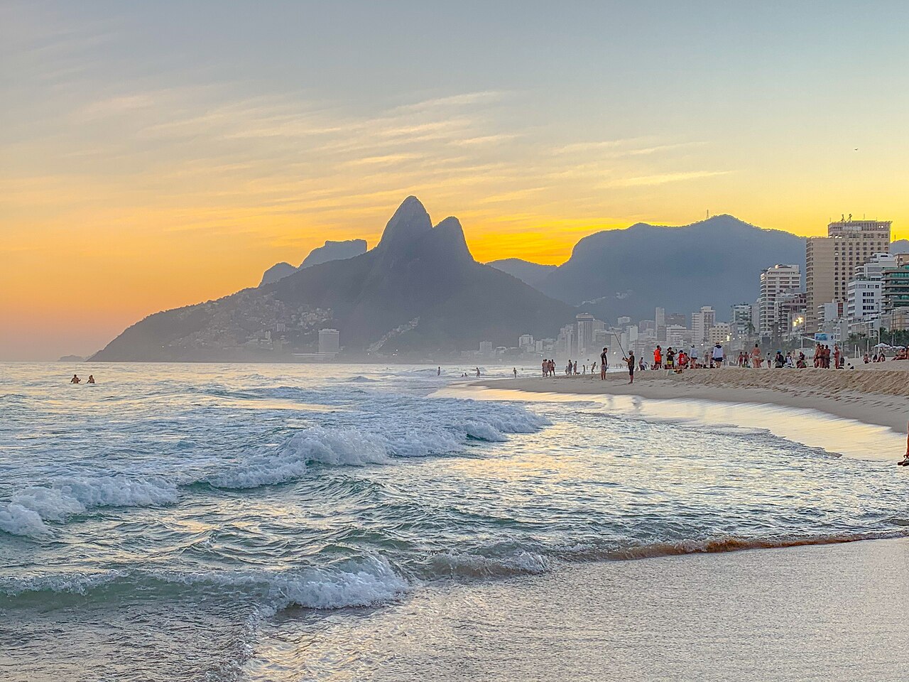 Waves roll onto a sandy beach beneath twin rocky peaks and a yellow evening sky.