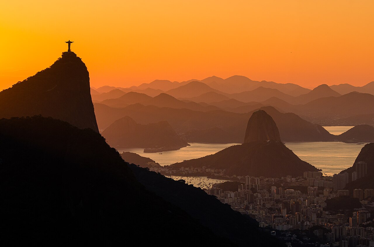 Dark mountain silhouettes, one topped by a statue, stand against an orange sky above a bay.