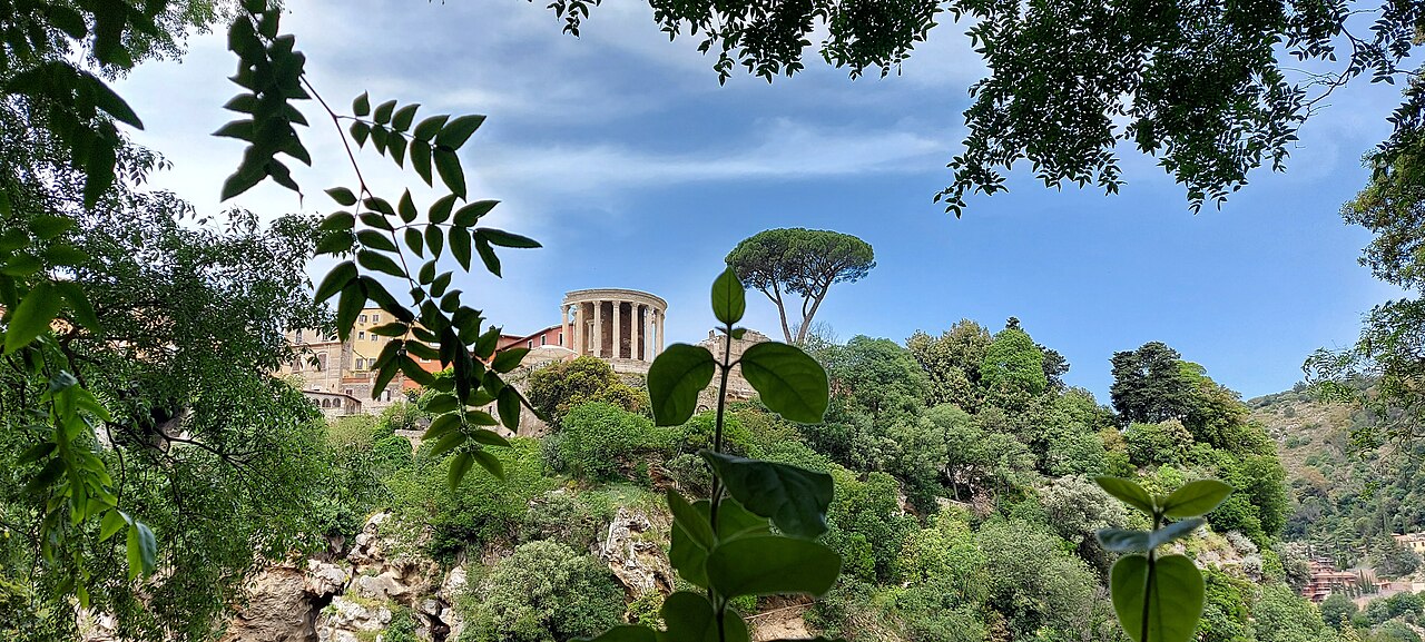 A small round colonnaded temple on a wooded hilltop, framed by leaves and an umbrella pine.