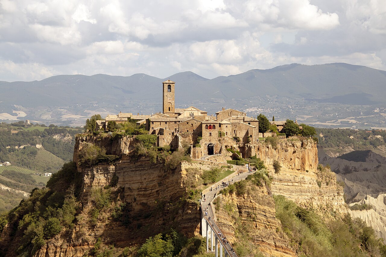 A cluster of stone houses and a bell tower on top of a steep eroded rock, with a narrow bridge climbing up to it.