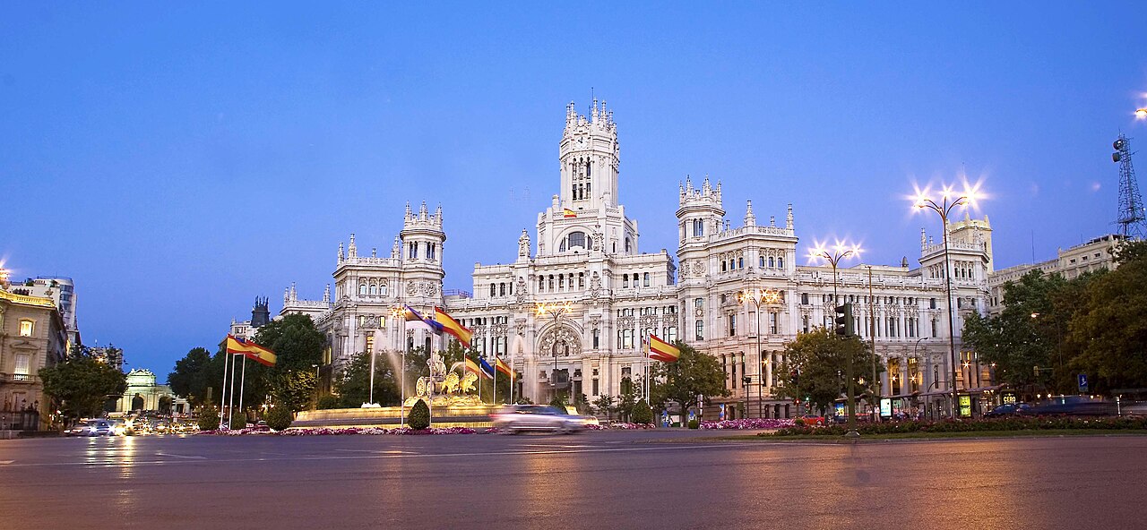 A large white palace with pointed towers, lit from below, behind a fountain and blurred evening traffic.