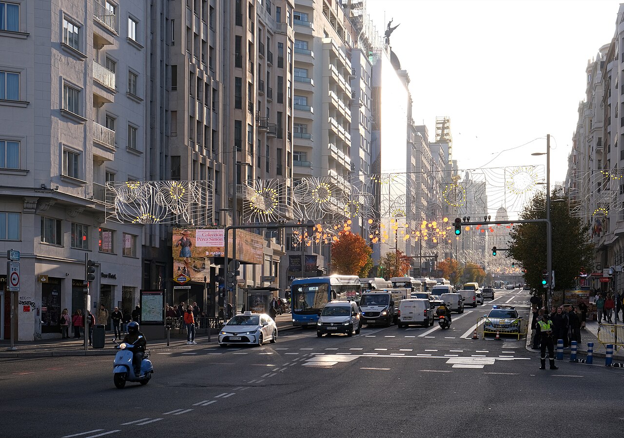 A wide city avenue with traffic and strings of lights overhead, lined by tall stone buildings.