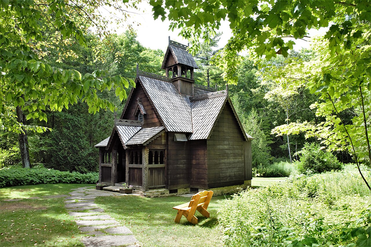 A small dark timber chapel with steep layered roofs and a spire stands in a green woodland clearing.