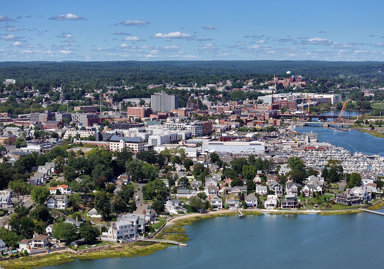 An aerial view of a small city with low brick buildings, tree-lined streets and a marina full of boats on a river.