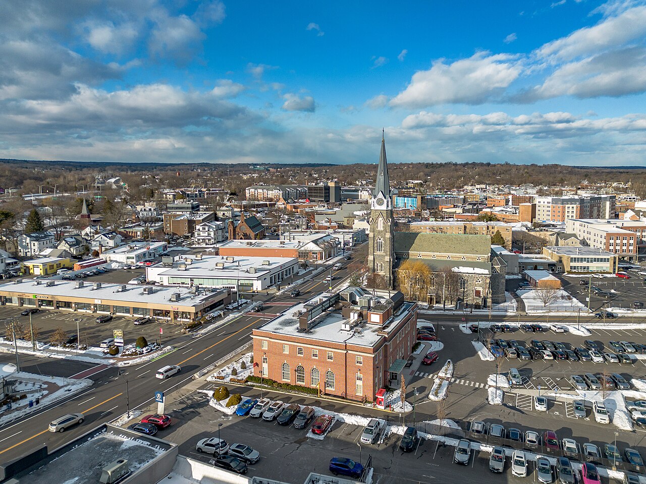 An aerial view over a town centre with a brick civic building, a tall church spire and bare trees under broken cloud.