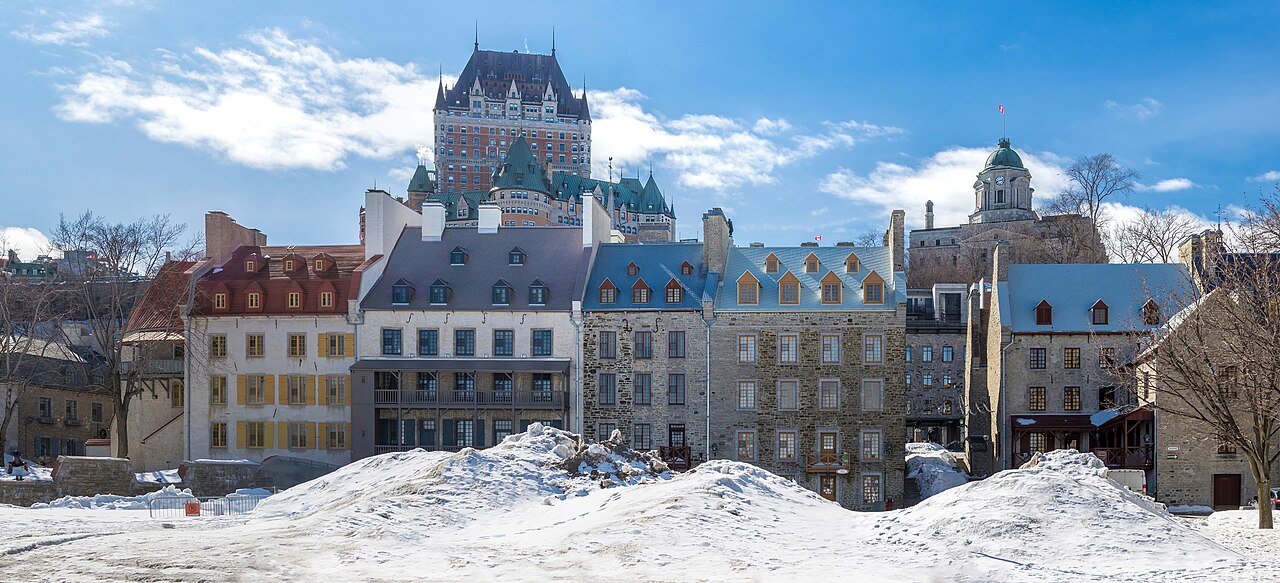 A row of old stone and brick houses behind a snow-covered square, with a large turreted hotel on the hill above.