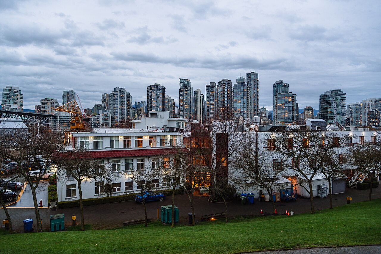 A line of glass residential towers rises behind low white waterfront buildings and bare trees on a grassy slope.