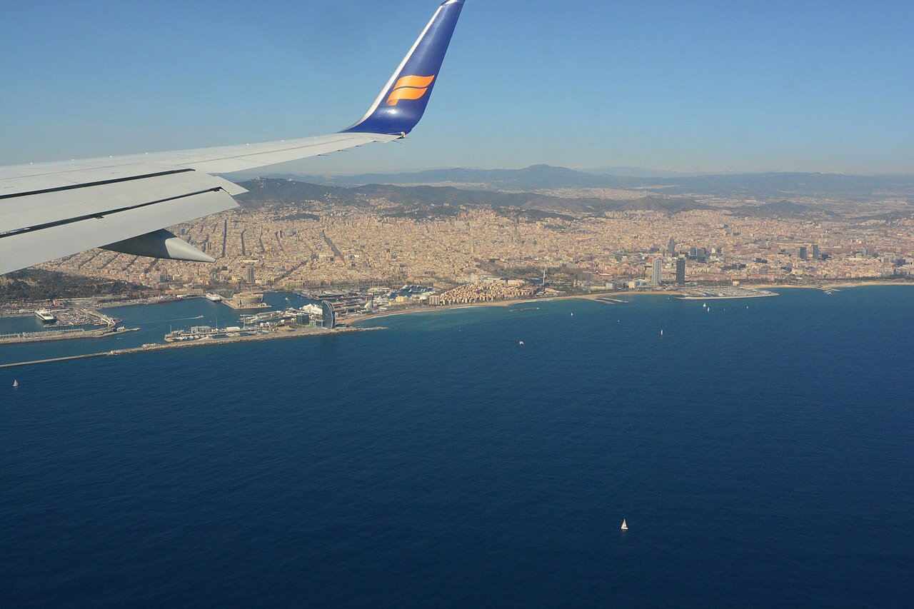 A city spreading from a curved shoreline up to low hills, seen over dark blue sea from an aircraft window with a wingtip in frame.
