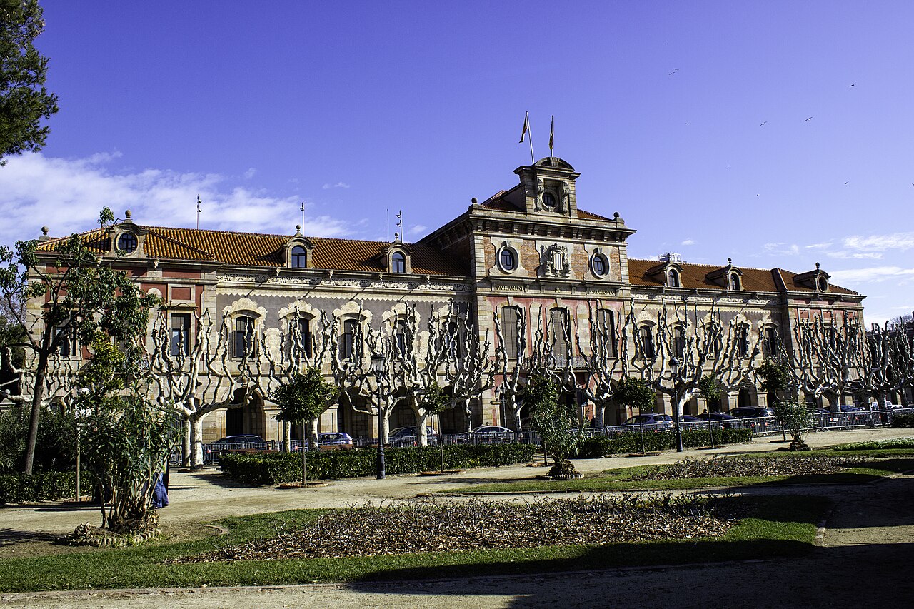 A long pale palace with arched ground-floor windows and a central flagpole, behind formal gardens and pruned trees.