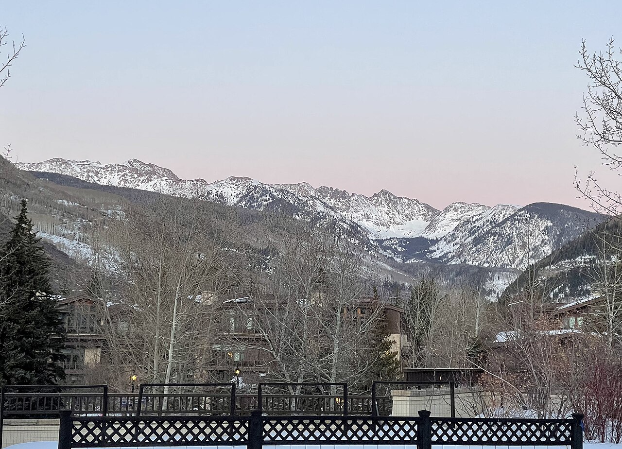 Snow-covered mountain peaks under a pink evening sky, above a valley of bare trees and rooftops.