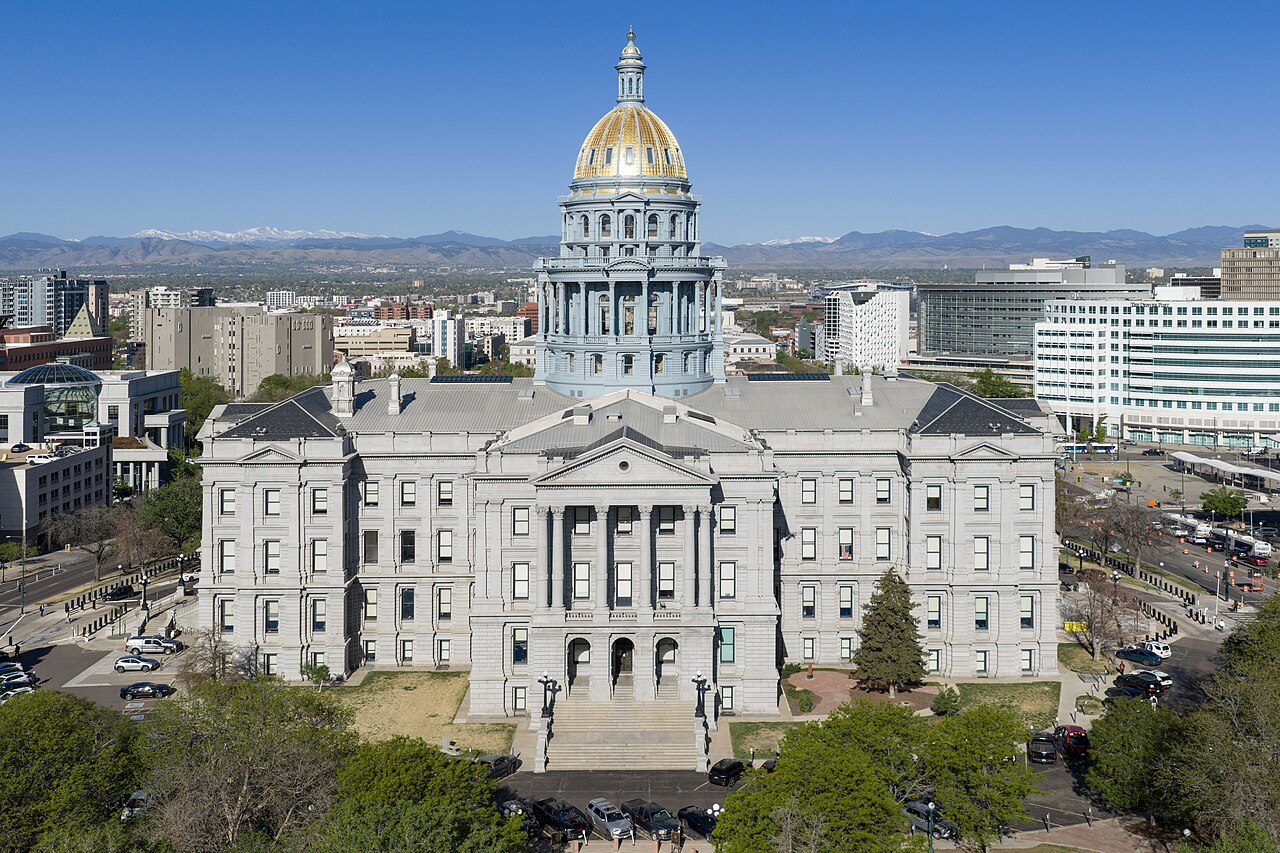 A grey granite capitol building with a gold dome seen from above, with the city and distant mountains behind it.