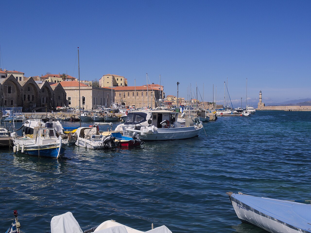 Small boats moored in a blue harbour in front of a row of old stone buildings, with a lighthouse at the far end of the breakwater.