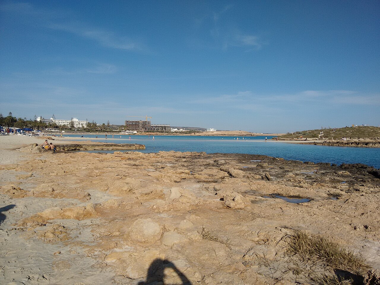 Flat pale rock in the foreground leads to a shallow turquoise bay and a strip of sand with low buildings beyond.
