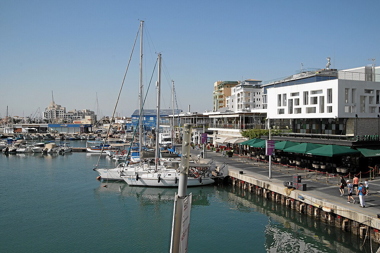 Sailing boats moored along a quay lined with white modern buildings and green cafe awnings.