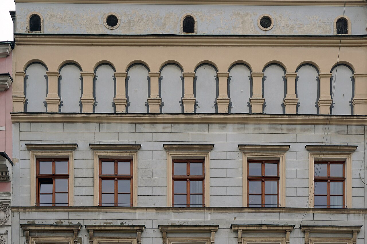 The upper floors of a pale stone townhouse with a row of arched blind arcades above tall rectangular windows.