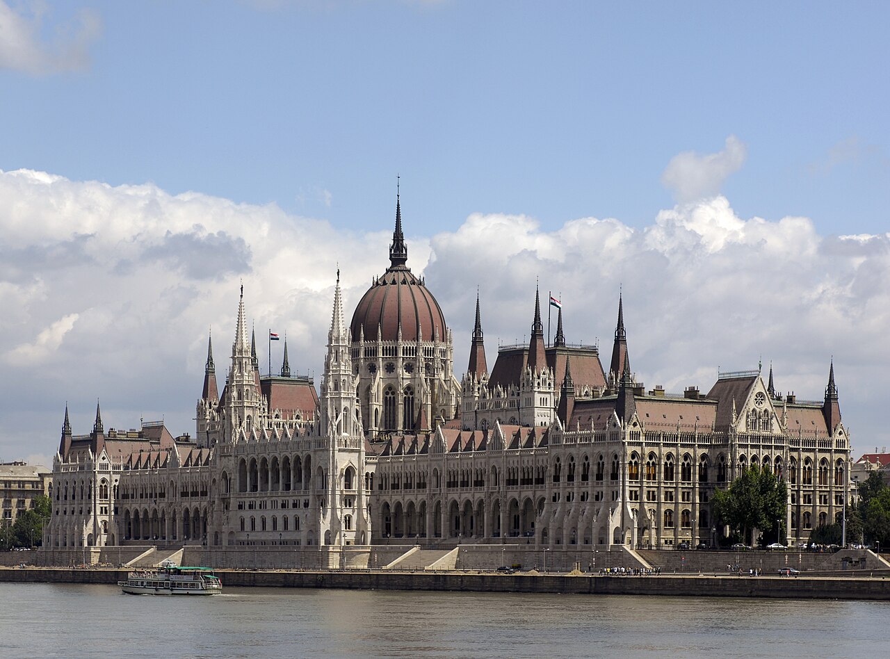 A long Gothic Revival building with a red central dome and rows of spires standing on a riverbank.