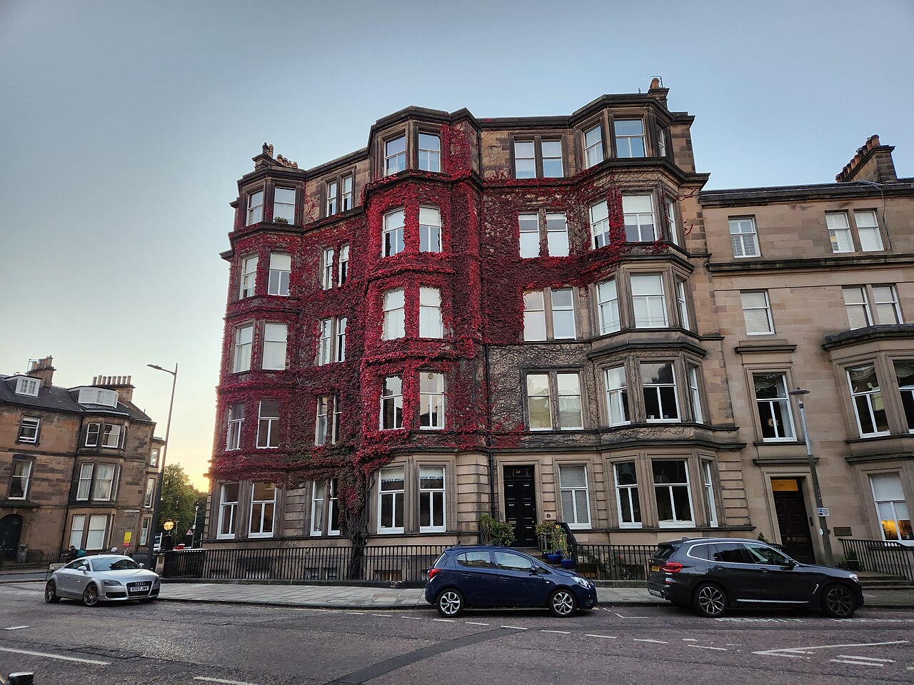 A terrace of four-storey stone townhouses with bay windows, one front covered in red creeper, with cars parked along the street.