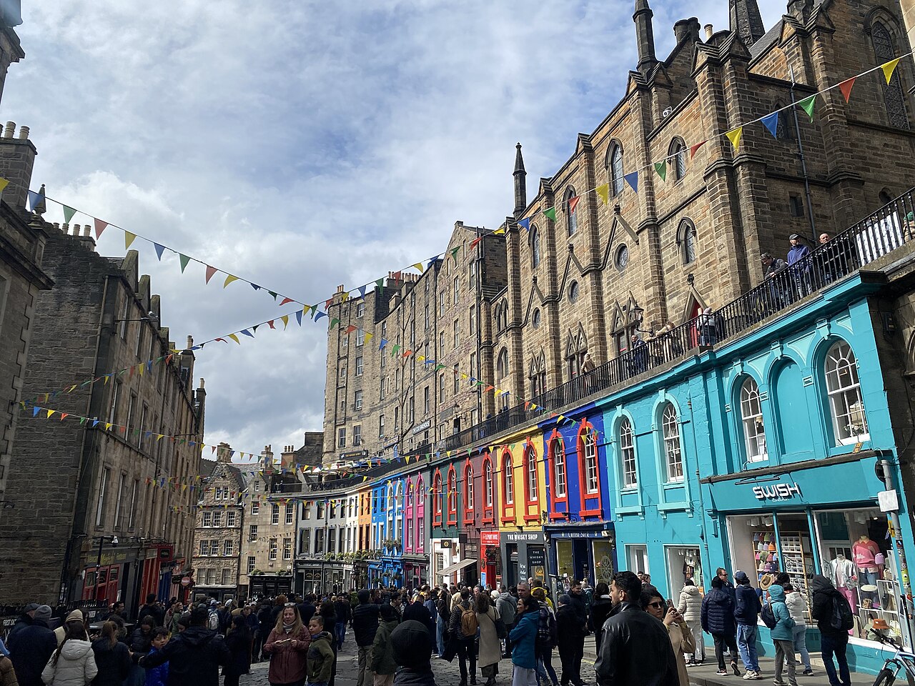 A curving street of tall stone tenements with brightly painted shopfronts, bunting overhead and a crowd of small figures below.